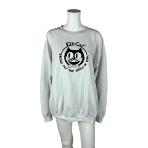 May include: A white sweatshirt with a black and white Kit-Cat logo. The logo features a cartoon cat with a bow tie and the text "Kit-Cat" and "Brings out the smile in you".