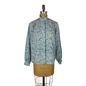 Op de afbeelding: Een lichtblauwe blouse met lange mouwen, een hoge kraag en een knoopsluiting. De blouse heeft een herhalend patroon van abstracte vormen in blauw, wit en roze. De blouse wordt getoond op een vintage mannequin.
