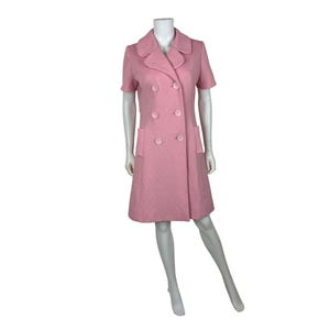 Puede incluir: Vestido rosa claro de doble botonadura con mangas cortas. El vestido presenta una tela texturizada, un cuello con muescas y dos bolsillos delanteros. El vestido se exhibe en un maniquí.