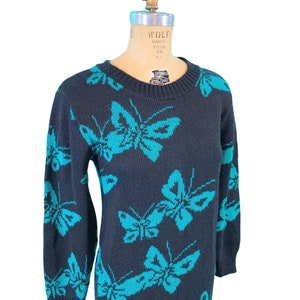 May include: A dark navy blue sweater dress featuring a repeating pattern of turquoise butterflies. The dress has a crew neck and long sleeves. The butterflies are a contrasting color, creating a striking visual effect. The dress is displayed on a mannequin.