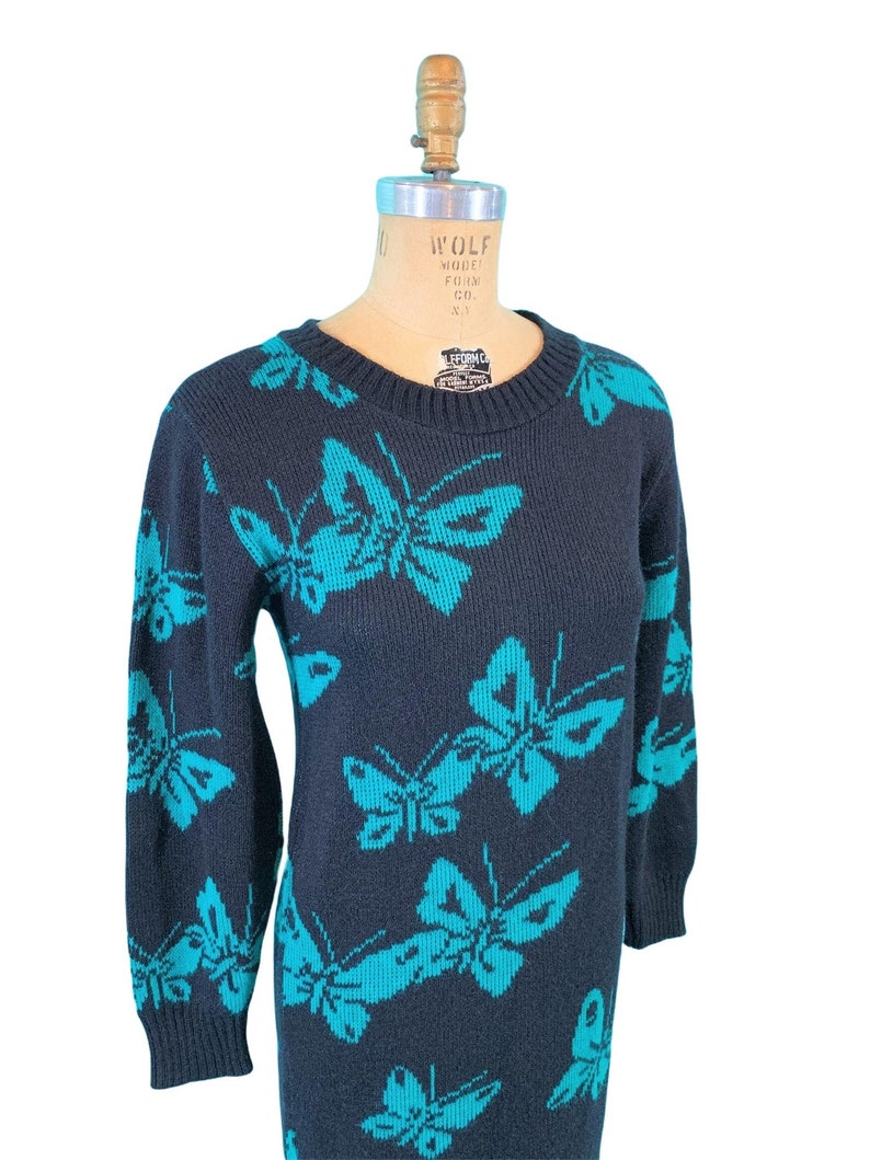 May include: A dark navy blue sweater dress featuring a repeating pattern of turquoise butterflies. The dress has a crew neck and long sleeves. The butterflies are a contrasting color, creating a striking visual effect. The dress is displayed on a mannequin.