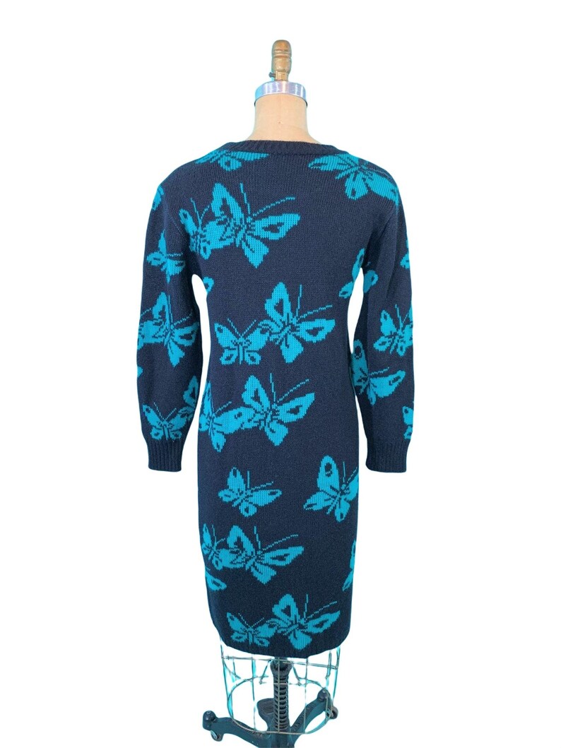 May include: A navy blue knit dress featuring a repeating pattern of turquoise butterflies. The long-sleeved dress is displayed on a mannequin, showcasing its form-fitting silhouette. The butterfly design adds a whimsical touch to the garment.