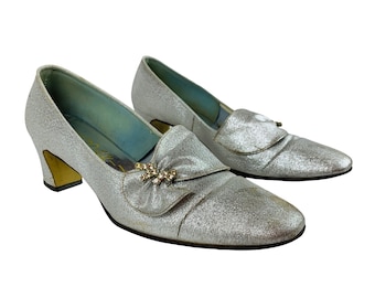 May include: A pair of silver glitter heeled shoes with a jeweled bow accent. The shoes have a low block heel and a pointed toe.