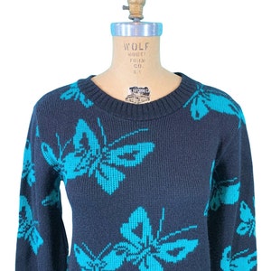May include: Navy blue sweater featuring a repeating pattern of turquoise butterflies. The sweater has a crew neck and long sleeves. The garment is displayed on a mannequin. The sweater is made of a knit fabric.