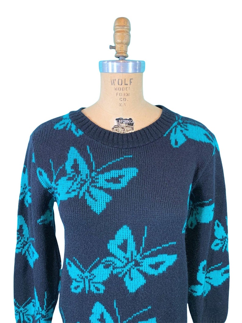 May include: Navy blue sweater featuring a repeating pattern of turquoise butterflies. The sweater has a crew neck and long sleeves. The garment is displayed on a mannequin. The sweater is made of a knit fabric.