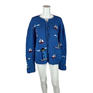 May include: A blue knitted cardigan with a sailor theme. The cardigan features a lighthouse, sailboats, and clouds embroidered on the front. The cardigan has a tie closure at the neck.