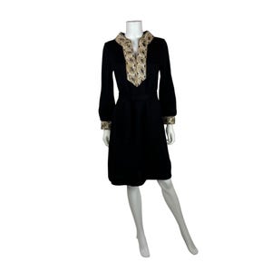 May include: A black dress with long sleeves and a V-neckline. The neckline and cuffs are adorned with a decorative gold and beige pattern. The dress is belted at the waist and falls to the knees.