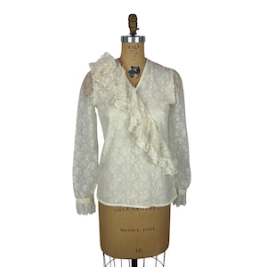May include: Ivory lace blouse with long sleeves and a ruffled asymmetrical neckline. The blouse is displayed on a vintage dress form. The sleeves have a floral lace pattern and the cuffs are trimmed with lace.