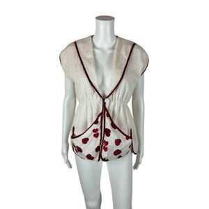 May include: Cream-colored vintage vest with a V-neck and cap sleeves. The vest features dark red trim and a heart-patterned panel at the bottom. A single white button fastens the front. The vest is displayed on a mannequin.