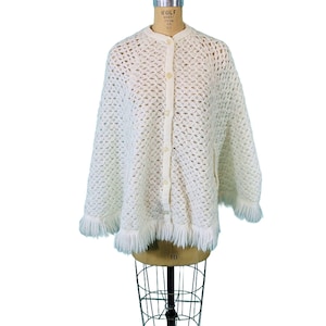 May include: White crocheted poncho with a fringe trim and button closure.
