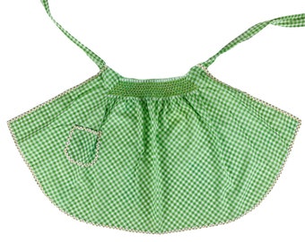 May include: A green gingham apron with a white trim and a small pocket. The apron has a gathered neckline and adjustable straps.