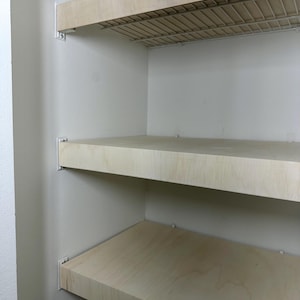 May include: A built-in shelving unit with three wooden shelves and a wire rack shelf. The shelves are light-colored and mounted on white brackets against a white wall. The unit is empty.