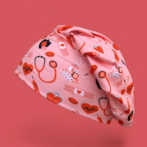 May include: A pink beanie hat with a repeating pattern of medical symbols, including stethoscopes, hearts, bandages, and syringes in red and white. The hat is made of a soft, flexible material.