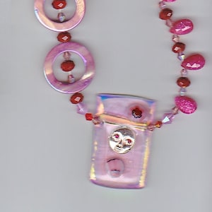 May include: A pink and red beaded necklace with a pink and silver pendant featuring a face. The pendant is rectangular and has a iridescent finish. The necklace has a silver clasp.