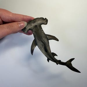 May include: A flat, cutout-style hammerhead shark figure featuring a dark, charcoal-gray finish with subtle shading and texture. The shark is depicted in a swimming pose with its distinct wide head, pectoral fins, and a forked tail fin.