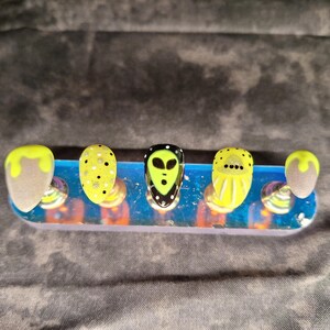 May include: Five nail tips with different designs. The tips are arranged on a blue and white display stand. The designs include a green alien face, yellow polka dots, and a solid yellow color.