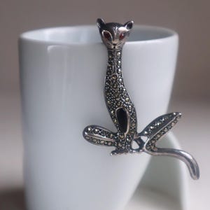 May include: A silver cat brooch with marcasite detailing and red gemstone eyes, attached to a white ceramic mug. The cat is in a seated pose with its tail curled. The brooch is a decorative item.