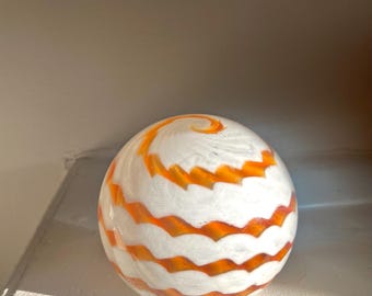 May include: A decorative glass sphere with a white base and orange, wavy stripes. The sphere has a spiral design at the top. The glass is smooth and reflective, and the orange stripes are evenly spaced around the sphere.