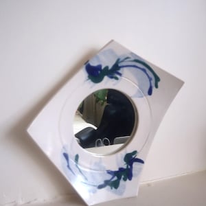 May include: A white, decorative mirror with a circular centre and an angled frame. The frame features blue and green floral patterns. The mirror is set against a white wall, offering a clean and artistic appearance.