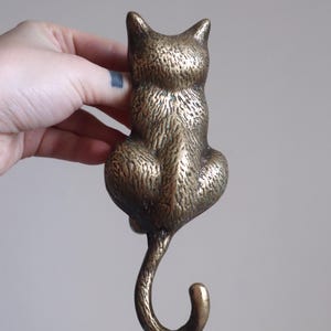 May include: Bronze cat-shaped hook, viewed from the back. The hook has a textured surface and a curled tail. The cat is in a sitting position, with its ears perked up. The hook is approximately 4 inches tall.
