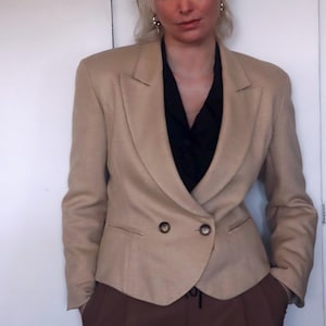 Könnte beinhalten: Beigefarbener Blazer mit Doppelreihverschluss, kombiniert mit brauner Hose und schwarzem Oberteil. Der Blazer hat ein klassisches Revers und zwei sichtbare Knöpfe. Das Outfit hat eine neutrale Farbpalette.