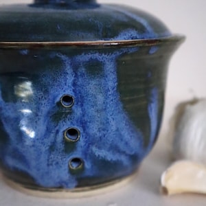 May include: A ceramic garlic keeper with a lid, glazed in blue and green hues. It features three ventilation holes. A clove of garlic is visible in the background. The keeper is a handmade item.
