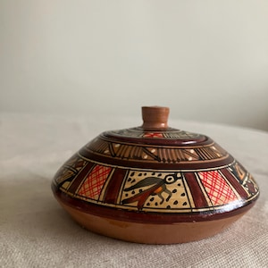 May include: A small, round, terracotta pottery jar with a lid. The jar is decorated with geometric patterns in red, brown, and white. The lid has a small knob on top.
