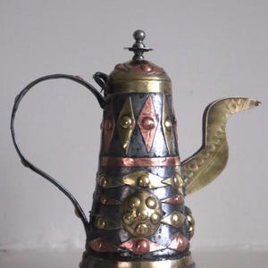 May include: An ornate metal teapot with a decorative design. The teapot features a combination of silver, copper, and brass elements, including diamond and circular patterns. It has a curved handle and a spout, and stands on three small legs.