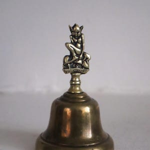 May include: A brass bell with a seated pixie figurine on top. The figurine has pointed ears and is perched on a small platform with the text "DARTMOOR PIXIE". The bell has a classic shape and a polished finish.