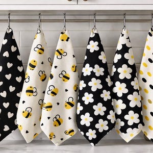 Pode incluir: Seis panos de cozinha estampados pendurados numa barra de metal. Os padrões incluem corações brancos em preto, abelhas em creme, flores brancas em preto e bolinhas amarelas e pretas em creme. Os panos estão num ambiente de cozinha.