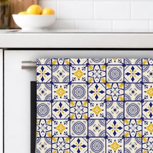 May include: A decorative kitchen towel with a repeating pattern of blue, yellow, and white tile designs. The towel is hanging on a white oven door. The tile pattern features geometric shapes and floral motifs.