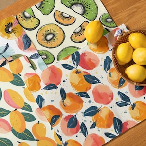 May include: A collection of colorful kitchen textiles featuring fruit designs. The textiles include placemats and a runner with patterns of kiwi, oranges, and lemons. A woven basket holds several fresh lemons, adding a vibrant touch to the scene.