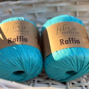 Può includere: Due gomitoli di rafia blu turchese. Il filo è etichettato "fibra natura" e "Raffia".