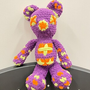 Puede incluir: Oso de peluche de crochet morado con detalles en forma de estrella amarilla y naranja. El oso está sentado en una silla negra.