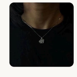 May include: A silver-toned necklace featuring a delicate chain and a small, open-work heart-shaped pendant. The pendant has an intricate, swirling design within the heart frame, resting against a dark, solid-coloured fabric background.