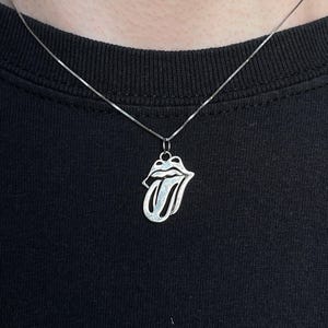 Könnte beinhalten: Eine silberne Halskette mit einem Anhänger, der das ikonische Zungen- und Lippen-Logo der Rolling Stones zeigt. Der Anhänger hängt an einer feinen Silberkette. Die Halskette wird vor einem schwarzen Hintergrund präsentiert.