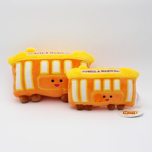 May include: Two plush orange cable car toys with yellow roofs and white window frames. The text "POWELL & MASON Sts." is printed on the roof of each toy. Each toy has a smiling face and brown wheels.