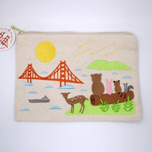 May include: Beige canvas zippered pouch with embroidered design. Features the Golden Gate Bridge, sun, deer, bears, rabbit, squirrel, and a log. The design is in orange, yellow, brown, pink, green, and blue. A small gold tag is attached to the zipper.