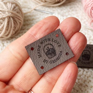 Peut inclure: Une étiquette carrée en similicuir marron foncé à la surface texturée, dotée de quatre trous aux coins pour la couture. L'étiquette est gaufrée avec un panier de laine et des aiguilles à tricoter, entouré du texte "MADE WITH LOVE BY GEORGIA".