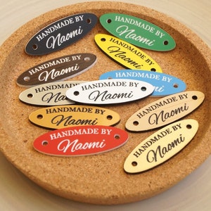 Könnte beinhalten: Eine Sammlung ovaler Holzetiketten mit dem Text "Handmade by Naomi" in schwarzer Schrift auf verschiedenen farbigen Hintergründen. Die Etiketten haben zwei Löcher für Schnur oder Band.