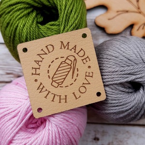 Peut inclure: Une étiquette carrée en cuir beige avec les mots "HAND MADE WITH LOVE" entourant un motif de bobine de fil et d'aiguille. L'étiquette est entourée de pelotes de laine vertes, roses et grises, suggérant un thème artisanal.
