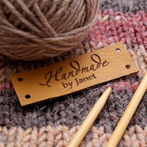 Könnte beinhalten: Ein rechteckiges, hellbraunes Kunstleder-Kleidungsetikett mit der Gravur Handmade by Janet in einer dunklen Schreibschrift. Das Etikett hat vier kleine, vorgestanzte Löcher an den Ecken und liegt auf einem strukturierten Strickstoff mit Garn und Nadeln.