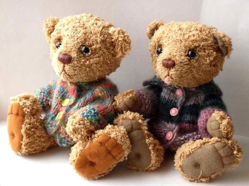 May include: Two brown teddy bears wearing knitted sweaters. The bear on the left is wearing a multi-colored sweater with a yellow button. The bear on the right is wearing a purple and black striped sweater with pink buttons.