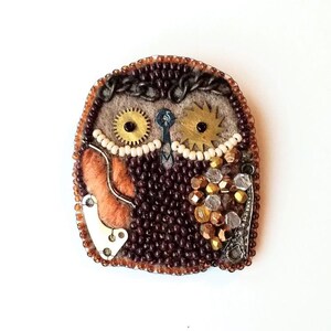 May include: A handcrafted owl brooch with a steampunk aesthetic. The owl is made of brown beads, felt, and metallic gears. It features orange, white, and silver accents, and is approximately 2.5 inches wide.