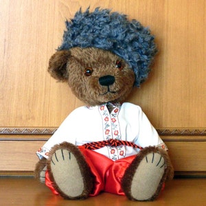 May include: A brown teddy bear wearing a white shirt with red floral embroidery, red pants, and a gray fur hat. The bear is sitting with its legs crossed. The background is a wooden cabinet.