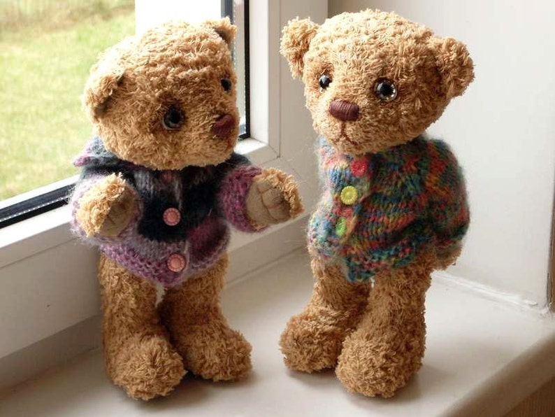 May include: Two brown teddy bears wearing knitted sweaters. One bear is wearing a purple and black sweater with pink buttons. The other bear is wearing a multi-colored sweater with yellow buttons.