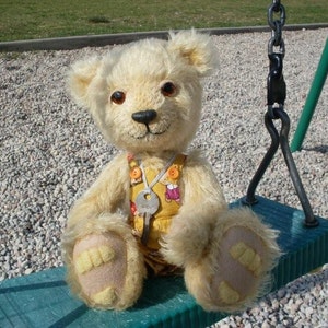 May include: A cream-colored teddy bear wearing a yellow and pink patterned overall with a key attached to a silver charm. The bear is sitting on a green swing set.