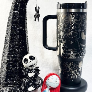Può includere: Una tazza da viaggio nera e argentata con un design che presenta personaggi del film "Nightmare Before Christmas". La tazza è appoggiata su un sottobicchiere nero. Ci sono due piccole statuette in ceramica di Jack Skellington e Sally del film. Un cappello da strega nero con un ragno d'argento appeso è sullo sfondo.