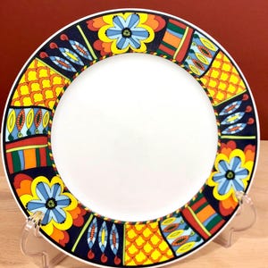 May include: A white ceramic plate with a colorful, geometric border. The border features a variety of shapes, including flowers, leaves, and abstract patterns. The colors in the border include red, orange, yellow, green, blue, and black.