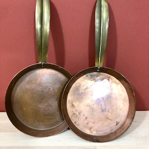 Può includere: Due padelle in rame vintage con manici in ottone. Le padelle sono rotonde e hanno una forma leggermente bombata. I manici sono lunghi e hanno un design semplice.